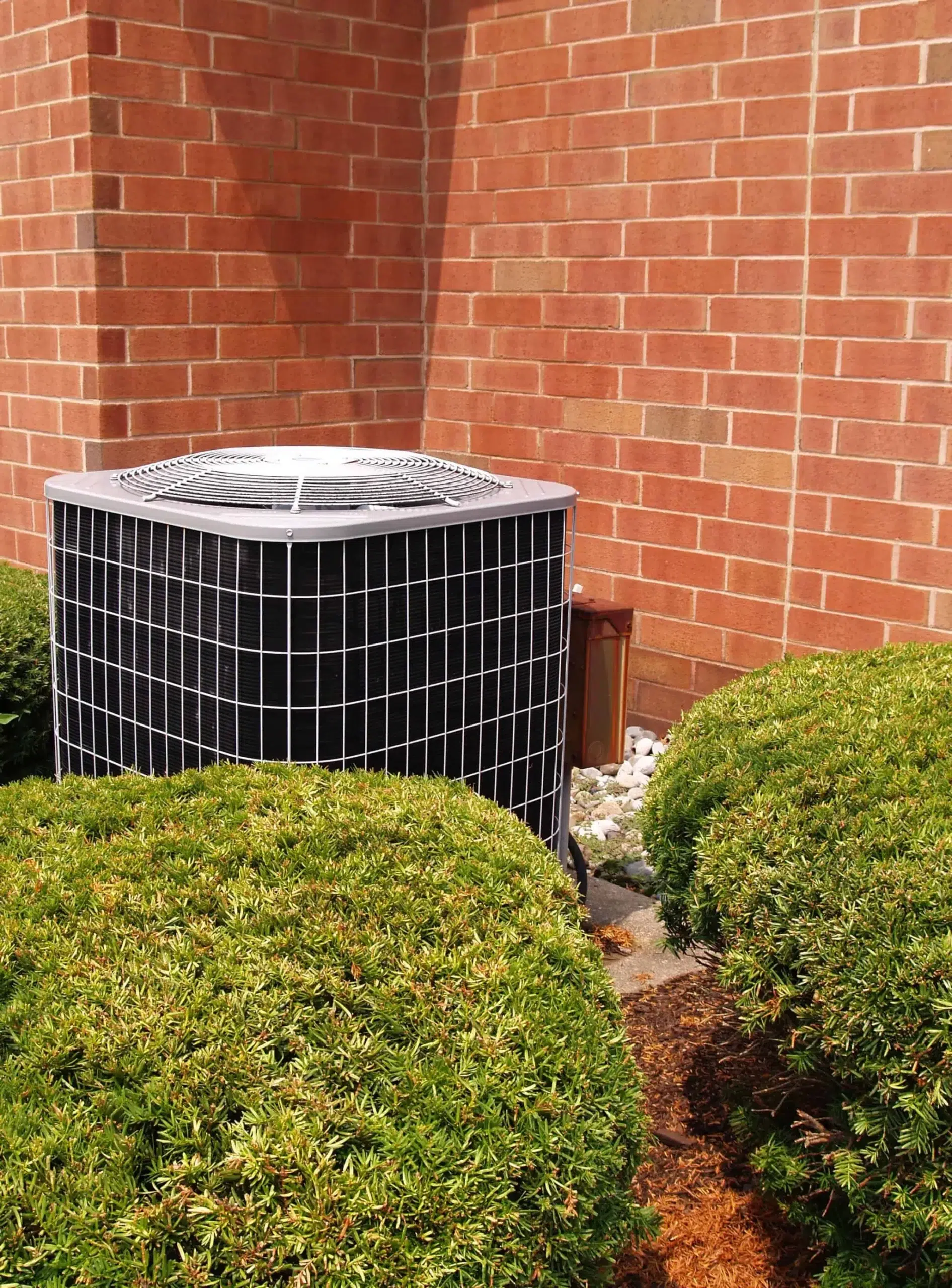 Outdoor air conditioning unit next to a brick wall, surrounded by green bushes and landscaping rocks.