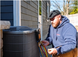 A technician in a blue hoodie inspects and works on an outdoor air conditioning unit next to a house.