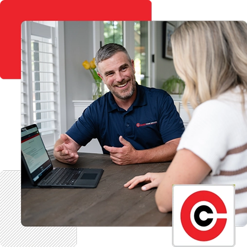 A man in a blue polo shirt discusses something with a woman at a table, with a laptop open in front of them. A red and black circular logo appears in the bottom right corner.