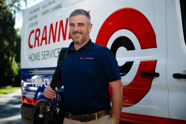 A smiling technician with a tool belt stands in front of a Cranny Home Services van, wearing a navy blue company polo shirt.