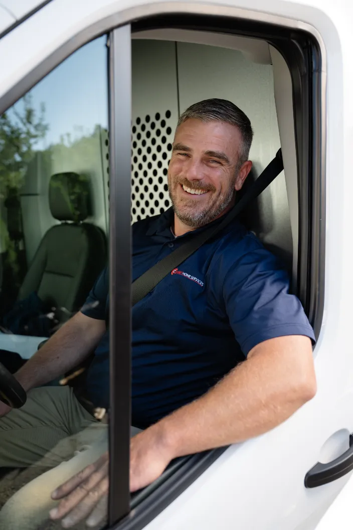 A man wearing a navy blue polo shirt sits in the driver’s seat of a white van, smiling at the camera with his seatbelt fastened.