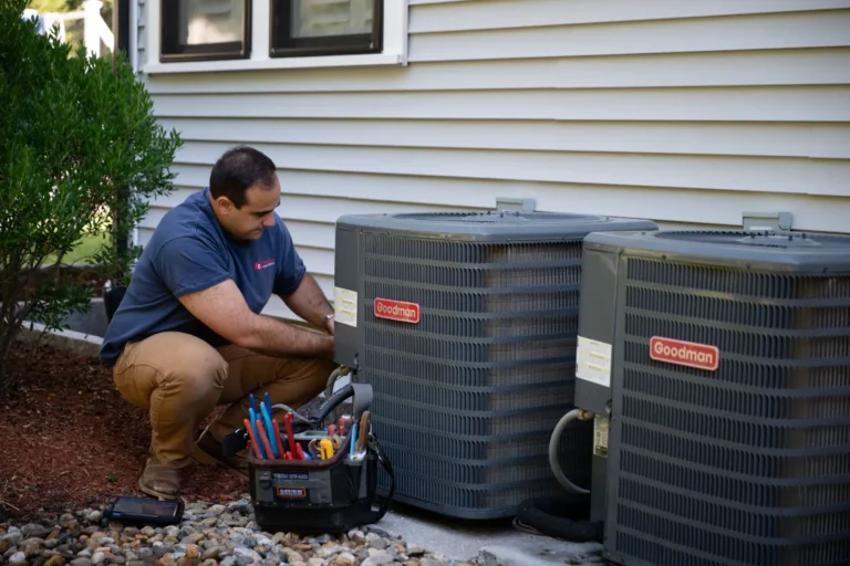 A technician kneels beside two outdoor Goodman HVAC units, using tools from a nearby tool bag to perform maintenance or repairs.