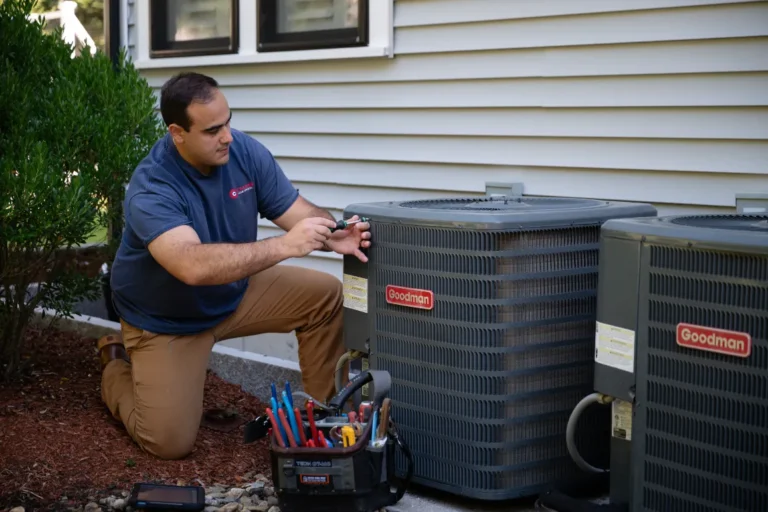 A technician kneels beside an outdoor Goodman air conditioning unit, using tools to perform maintenance or repairs. A tool bag is placed nearby.