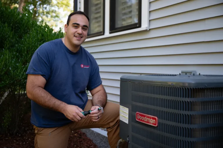 A person kneels next to a Goodman outdoor air conditioning unit, holding tools, beside a house with vinyl siding and shrubs.