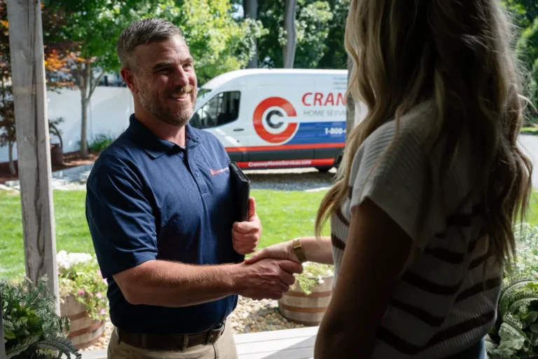 A technician in a blue polo shirt shakes hands with a woman at her doorstep, with a company van parked outside in the background.