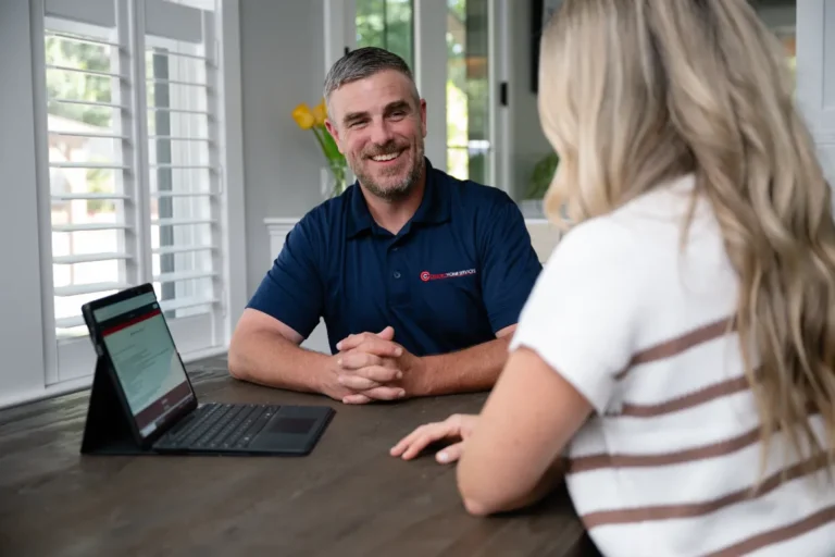 A man in a navy polo shirt sits at a table across from a woman, smiling, with a laptop open in front of them in a bright room.