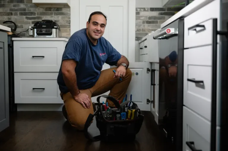 A man in a blue shirt kneels in a kitchen next to an open cabinet, with a tool bag full of tools on the floor.