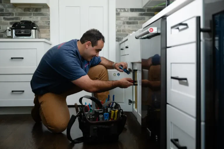 A man kneels on a kitchen floor using tools to work under a cabinet, with a tool bag placed nearby.