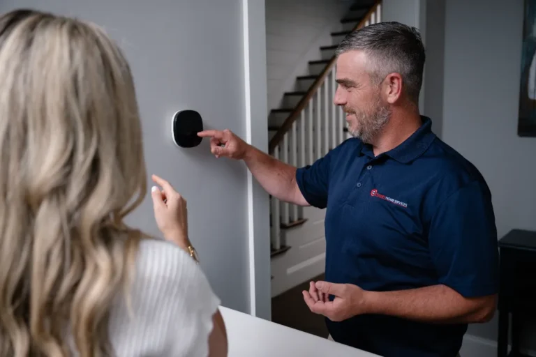 A man wearing a navy polo adjusts a wall-mounted thermostat while a woman stands nearby, inside a modern home.