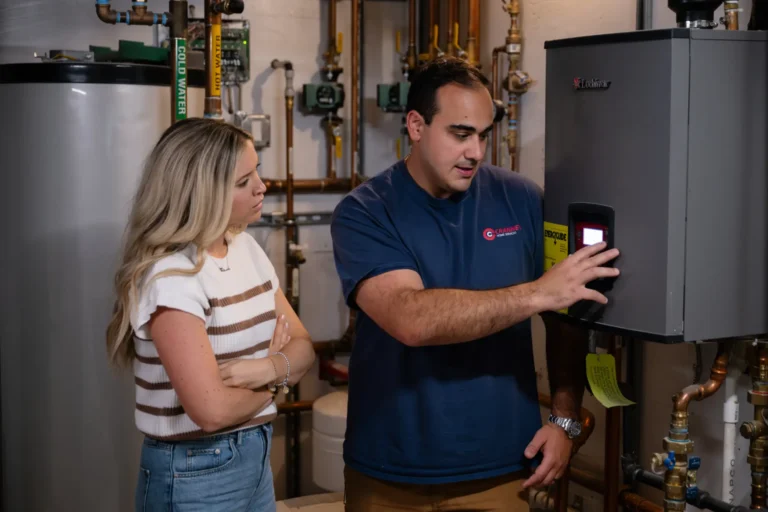 A man demonstrates controls on a wall-mounted heating unit to a woman in a utility room with visible pipes and equipment.