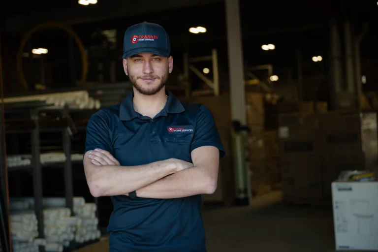 A man wearing a navy polo shirt and cap with a company logo stands with arms crossed in a warehouse setting.