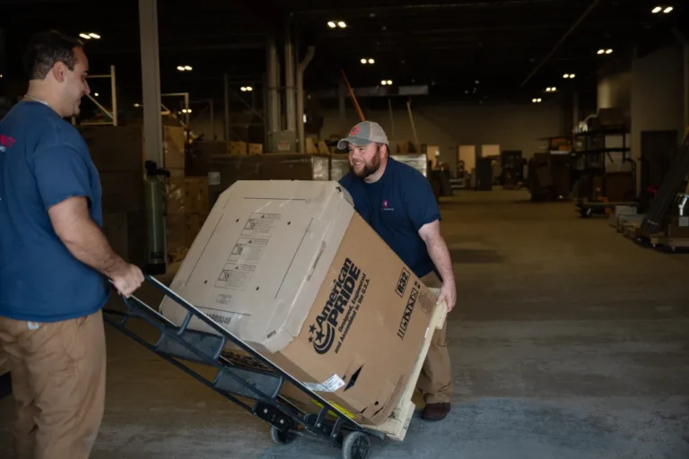 Two workers in a warehouse move a large cardboard box on a hand truck, with shelves and boxes visible in the background.