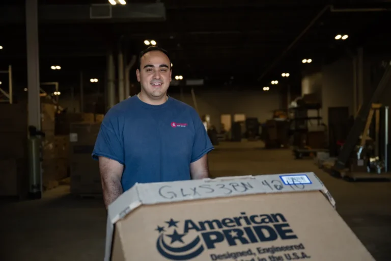 A man in a blue shirt stands in a warehouse, smiling, with a large cardboard box labeled "American Pride" in the foreground.
