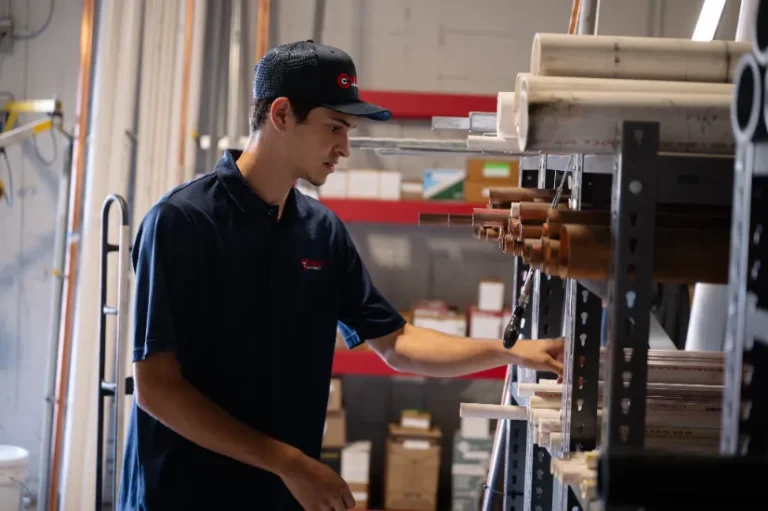 A person in a navy shirt and hat selects a pipe from a shelf stocked with various pipes in a workshop or storage area.