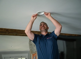 A man in a blue shirt installs or adjusts a circular fixture on a white ceiling inside a home.