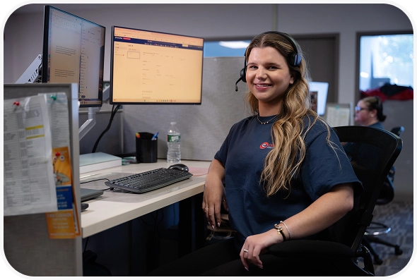 A woman wearing a headset sits at an office desk with dual monitors, smiling at the camera. Another person works in the background.