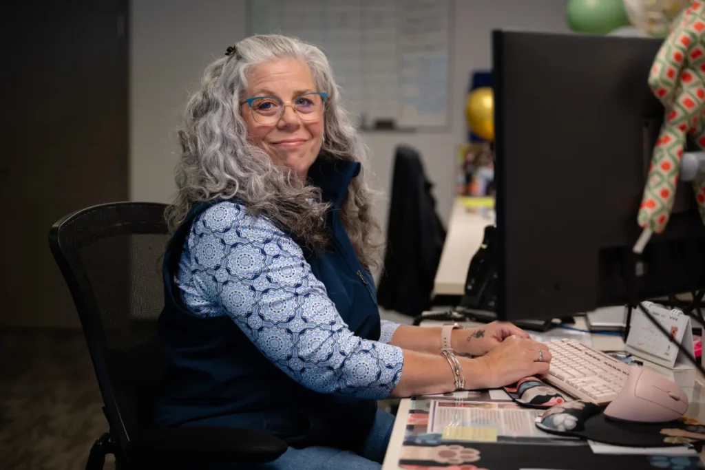 A woman with long gray hair and glasses sits at an office desk, typing on a keyboard and looking at the camera.