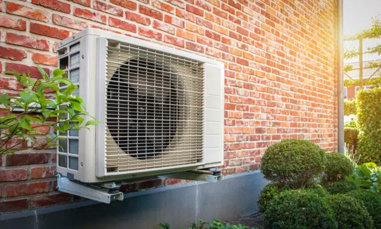 Outdoor air conditioning unit mounted on a red brick wall in Salem, surrounded by green shrubs and plants in sunlight.