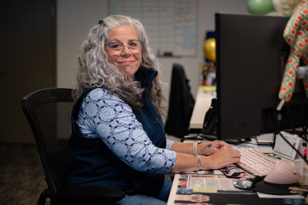 A friendly Cranney Home Services team member with gray hair and glasses types at her office desk, smiling warmly at the camera.