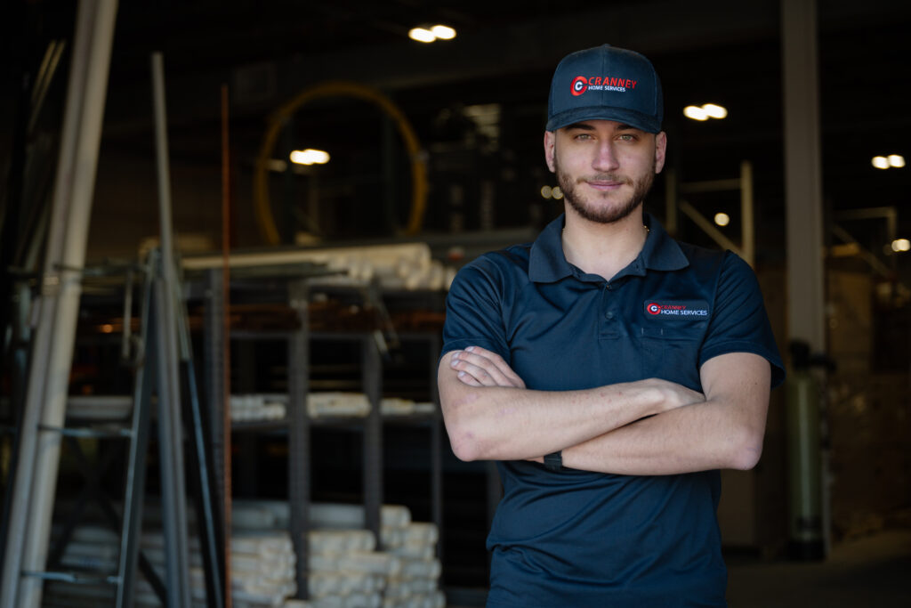 A Cranney Home Services team member in uniform stands confidently with arms crossed in a clean, organized warehouse facility.